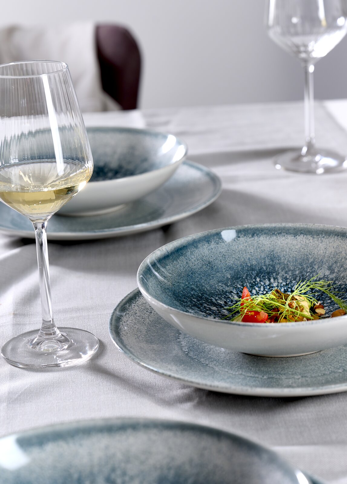Nahaufnahme eines gedeckten Tisches mit einem eleganten Tellerset in Blau- und Grautönen, darauf ein kunstvoll angerichtetes Gericht, im Hintergrund ein Glas Weißwein.