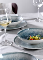Nahaufnahme eines gedeckten Tisches mit einem eleganten Tellerset in Blau- und Grautönen, darauf ein kunstvoll angerichtetes Gericht, im Hintergrund ein Glas Weißwein.