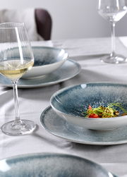 Nahaufnahme eines gedeckten Tisches mit einem eleganten Tellerset in Blau- und Grautönen, darauf ein kunstvoll angerichtetes Gericht, im Hintergrund ein Glas Weißwein.