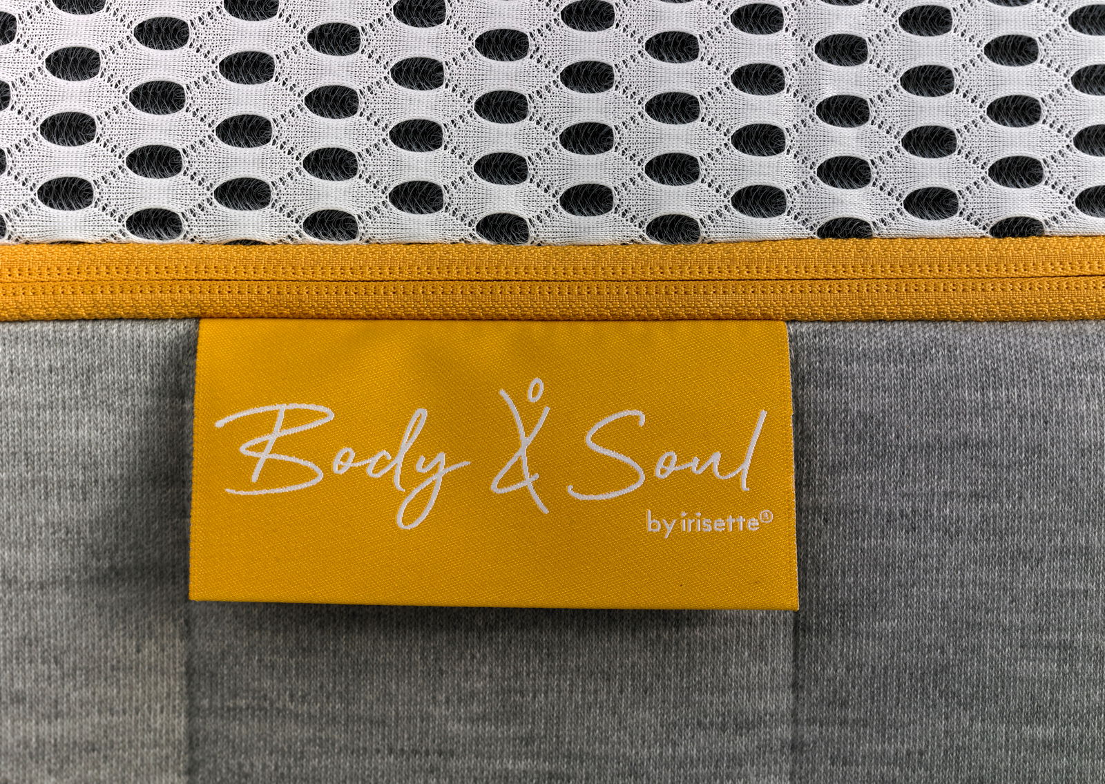 Nahaufnahme einer Matratze mit gelbem Etikett 'Body & Soul by irisette', grauem Stoff und weißem, gemustertem Obermaterial.
