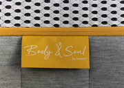 Nahaufnahme einer Matratze mit gelbem Etikett 'Body & Soul by irisette', grauem Stoff und weißem, gemustertem Obermaterial.