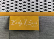 Nahaufnahme einer Matratze mit gelbem Etikett 'Body & Soul by irisette', grauem Stoff und weißem, gemustertem Obermaterial.