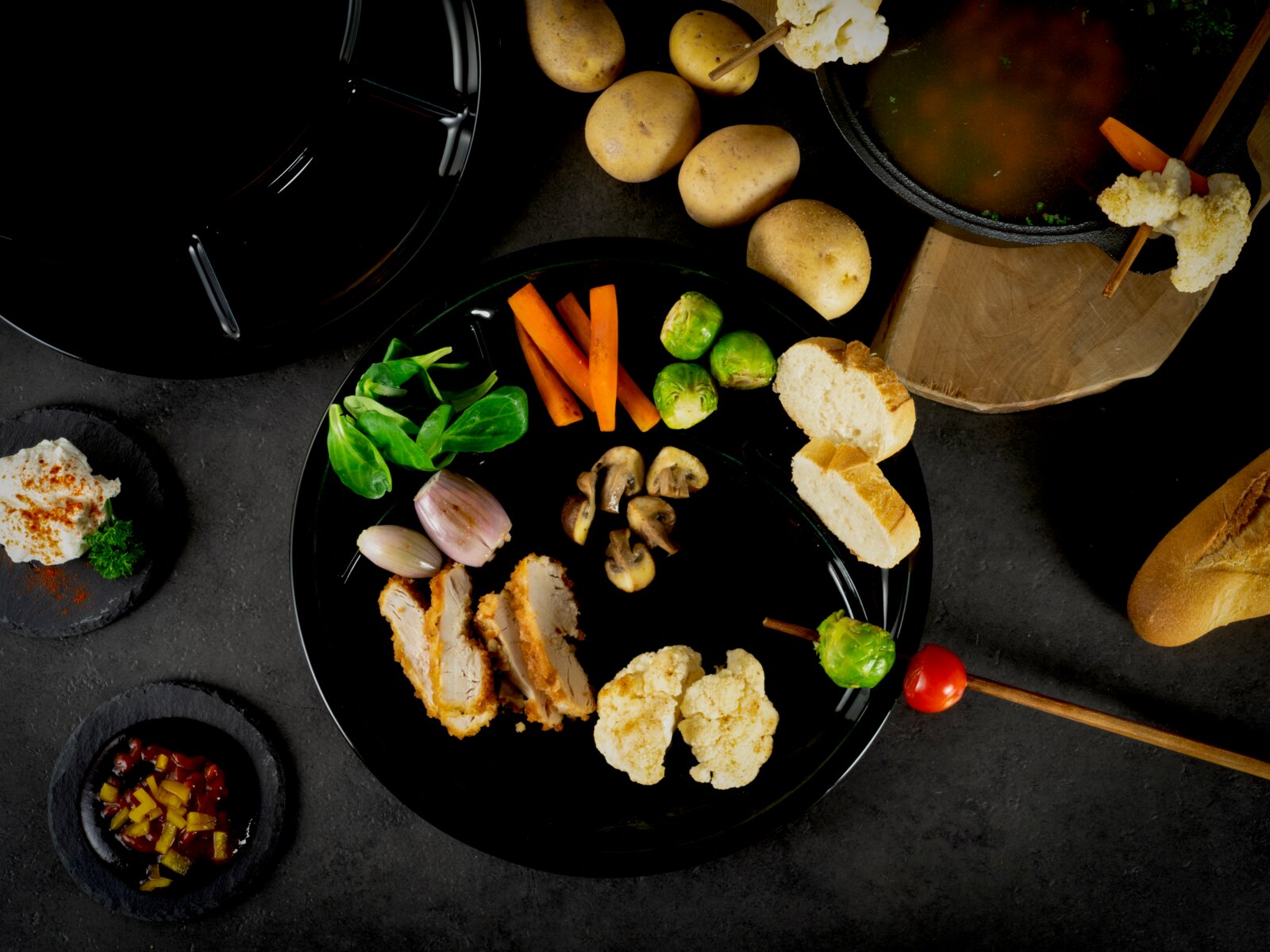 Draufsicht auf einen schwarzen Teller mit panierten Hähnchenstreifen, Blumenkohl, Champignons, Rosenkohl, Karottensticks, Feldsalat, Schalotten und Brotscheiben, daneben Kartoffeln und ein Topf Brühe auf dunklem Hintergrund