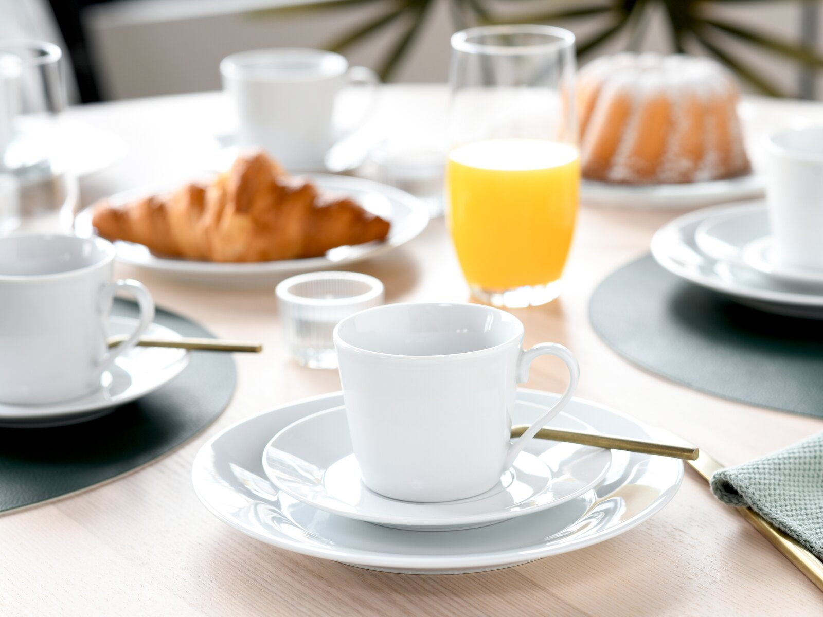 Schlichtes weißes Kaffeetassen-Set auf Untertasse und Teller auf gedecktem Frühstückstisch mit Croissant und Orangensaft, Perspektive: leicht von oben in Nahaufnahme.