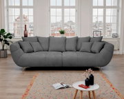 Großes graues Stoffsofa mit mehreren Kissen in einem hellen Wohnzimmer vor hohen Sprossenfenstern, frontale Ansicht auf das Produkt.
