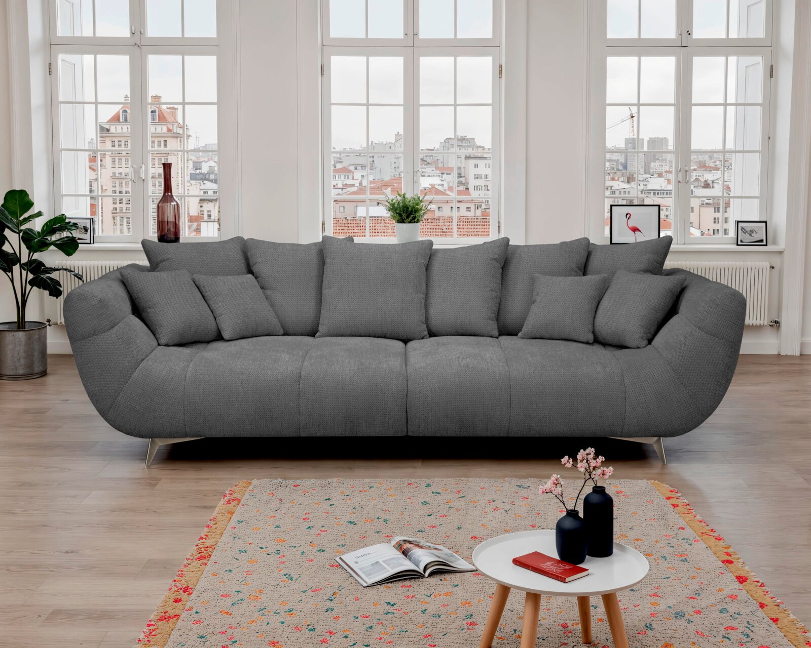 Großes graues Stoffsofa mit mehreren Kissen in einem hellen Wohnzimmer vor hohen Sprossenfenstern, frontale Ansicht auf das Produkt.