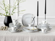Modernes weißes Porzellan-Geschirrset mit schwarzen und goldenen Linien (Teller, Schalen und Tassen) dekorativ auf einem gedeckten Tisch arrangiert, aufgenommen in Frontansicht auf Augenhöhe.