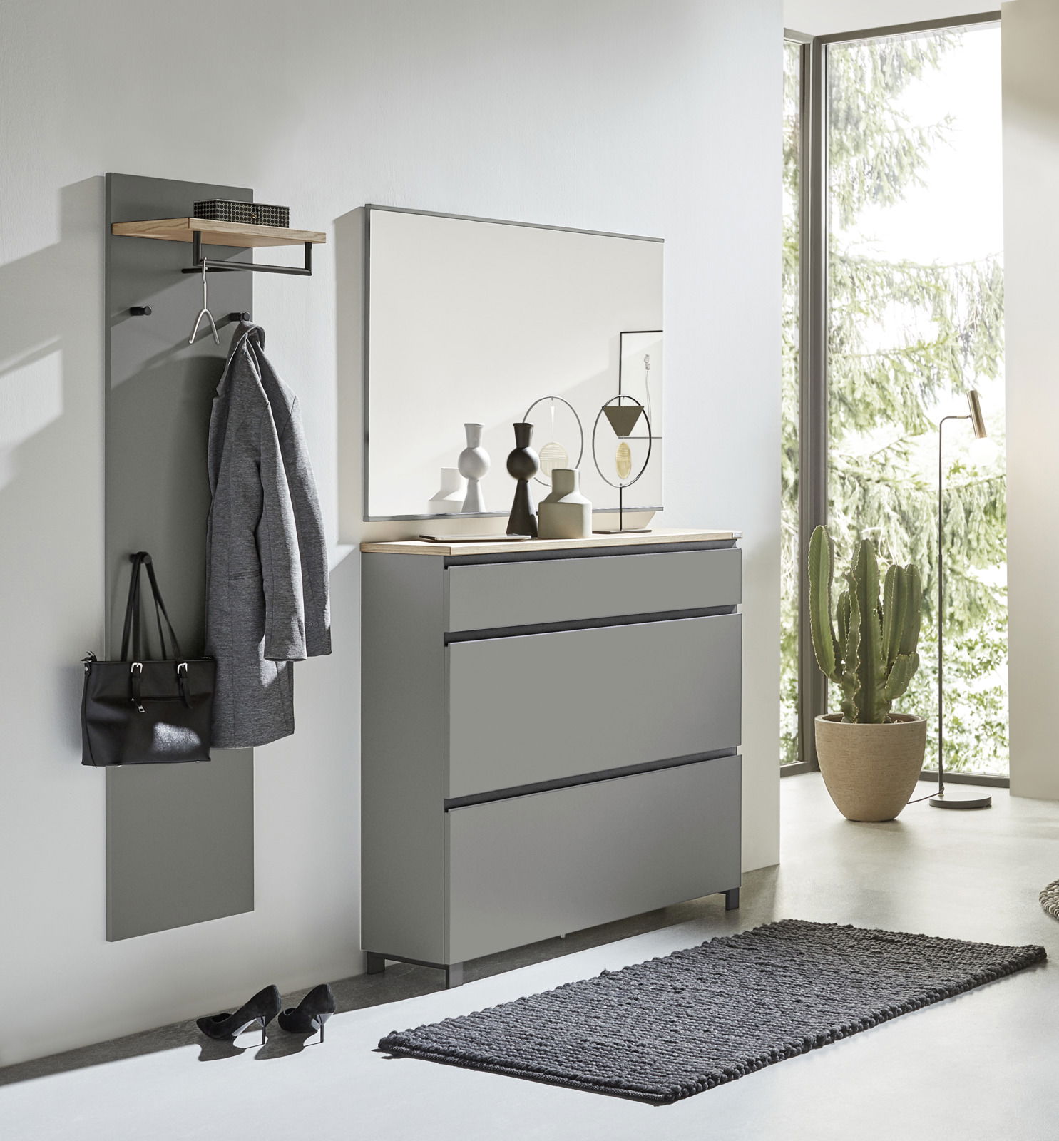 Modernes Garderoben-Set in Grau, bestehend aus einer Wandgarderobe mit Haken und Ablage, einem großen Spiegel und einer Kommode mit drei Schubladen. Perspektive: Frontalansicht in einem hellen Raum mit großen Fenstern.
