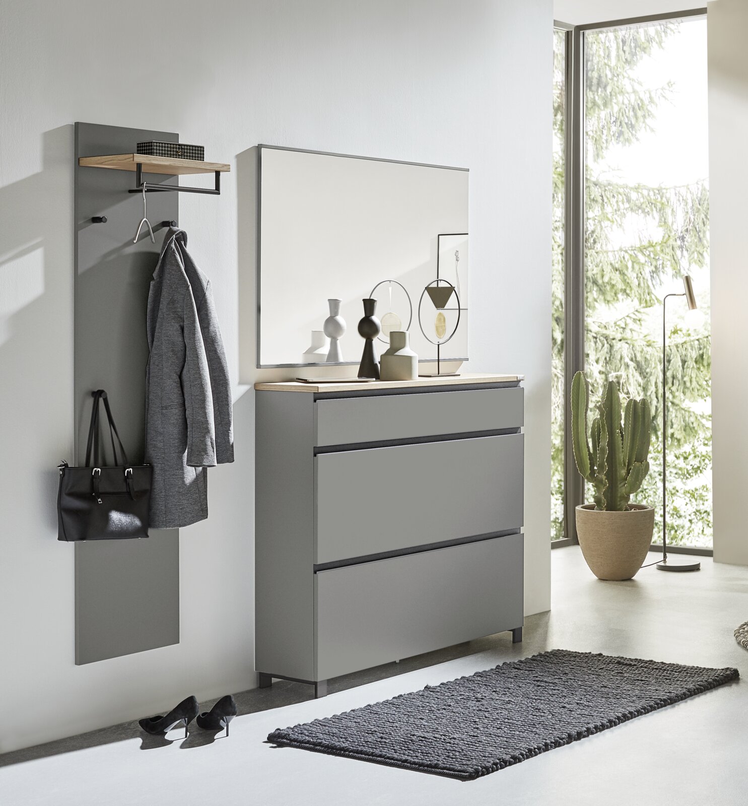Modernes Garderoben-Set in Grau, bestehend aus einer Wandgarderobe mit Haken und Ablage, einem großen Spiegel und einer Kommode mit drei Schubladen. Perspektive: Frontalansicht in einem hellen Raum mit großen Fenstern.