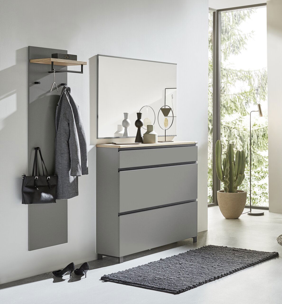 Dieter Knoll Garderoben-Set 3-tlg. SORRENT Modernes Garderoben-Set in Grau, bestehend aus einer Wandgarderobe mit Haken und Ablage, einem großen Spiegel und einer Kommode mit drei Schubladen. Perspektive: Frontalansicht in einem hellen Raum mit großen Fenstern.