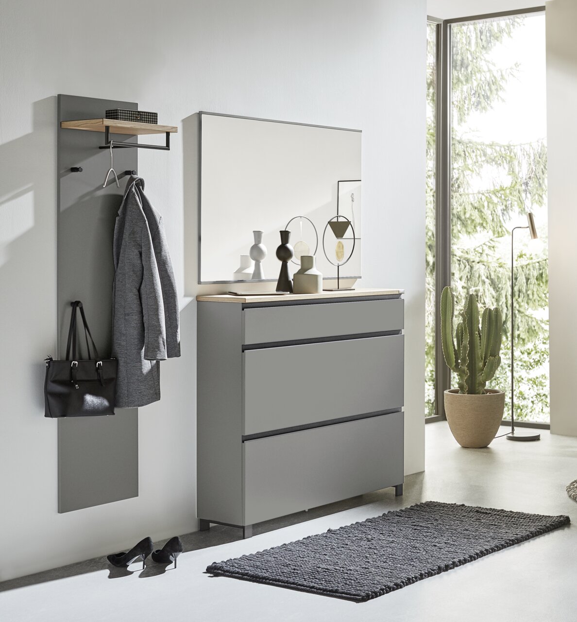 Dieter Knoll Garderoben-Set 3-tlg. SORRENT Modernes Garderoben-Set in Grau, bestehend aus einer Wandgarderobe mit Haken und Ablage, einem großen Spiegel und einer Kommode mit drei Schubladen. Perspektive: Frontalansicht in einem hellen Raum mit großen Fenstern.