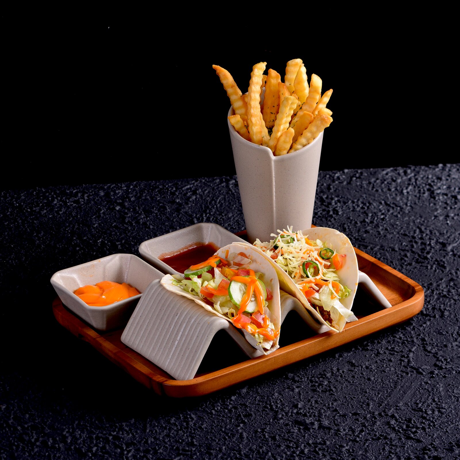 Schräg von vorn fotografiertes Serviertablett aus Holz mit weißen Keramikschalen: zwei gefüllte Tacos, Pommes frites im hohen Becher und zwei Dips auf dunklem Hintergrund.