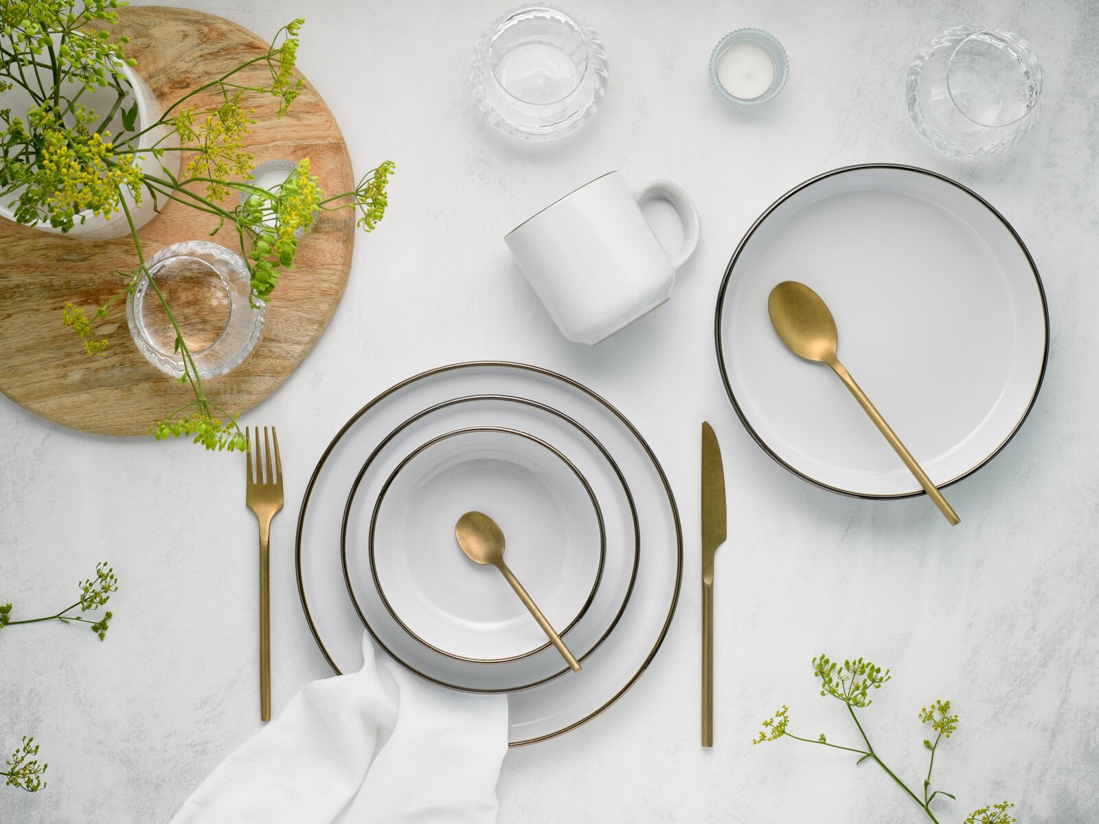 Draufsicht auf ein elegantes Tischgedeck mit weißen Tellern mit feinem Goldrand, goldfarbenem Besteck, weißer Tasse, Glas-Schälchen und grünen Blumen auf heller Steinoberfläche.