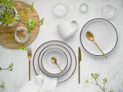 Draufsicht auf ein elegantes Tischgedeck mit weißen Tellern mit feinem Goldrand, goldfarbenem Besteck, weißer Tasse, Glas-Schälchen und grünen Blumen auf heller Steinoberfläche.