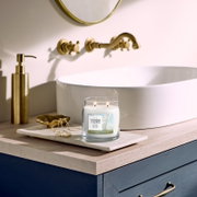 Yankee Candle Clean Cotton geurkaars met twee brandende lonten, vanuit vooraanzicht op een marmeren blad naast een elegante witte wastafel