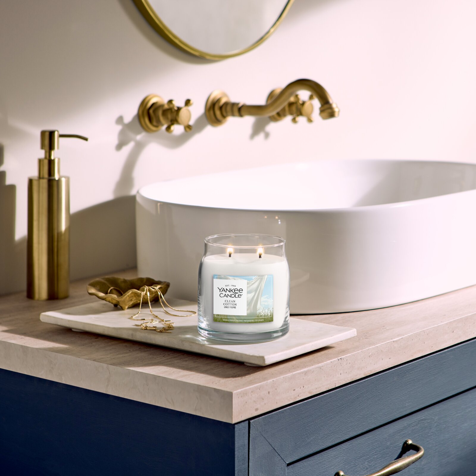 Yankee Candle Clean Cotton geurkaars met twee brandende lonten, vanuit vooraanzicht op een marmeren blad naast een elegante witte wastafel