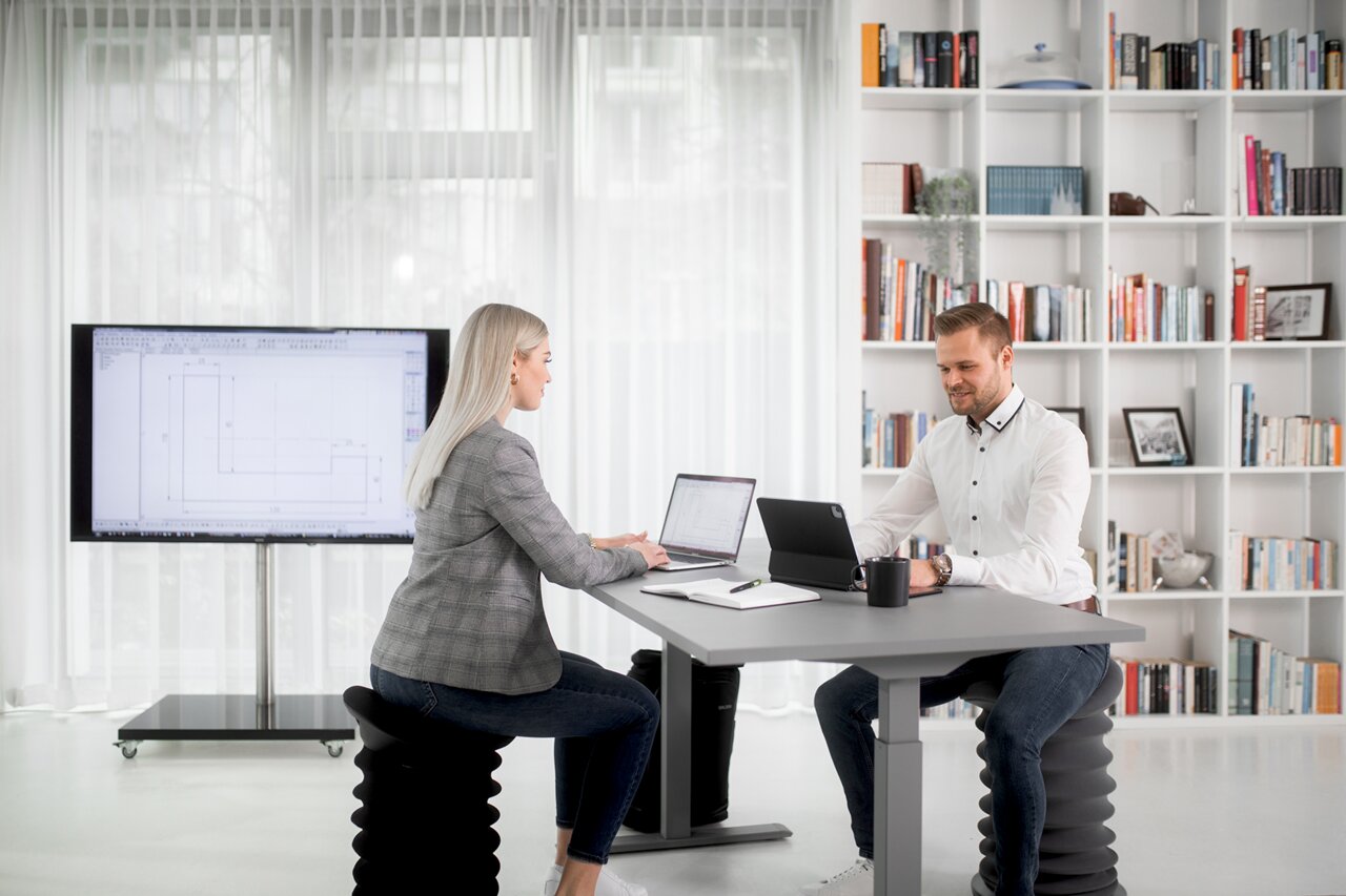 Zwei Personen arbeiten an einem höhenverstellbaren E-Schreibtisch in einem modernen Büro mit Bücherregal im Hintergrund, seitliche Perspektive.