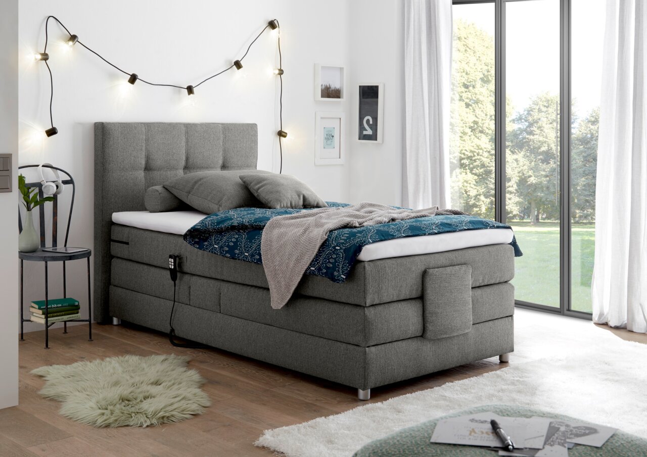 Esposa Boxspringbett MANOLO Graues Boxspringbett in einem modernen Schlafzimmer, seitliche Perspektive, mit dekorativen Kissen und Decken, umgeben von stilvollen Lichterketten und großen Fenstern.