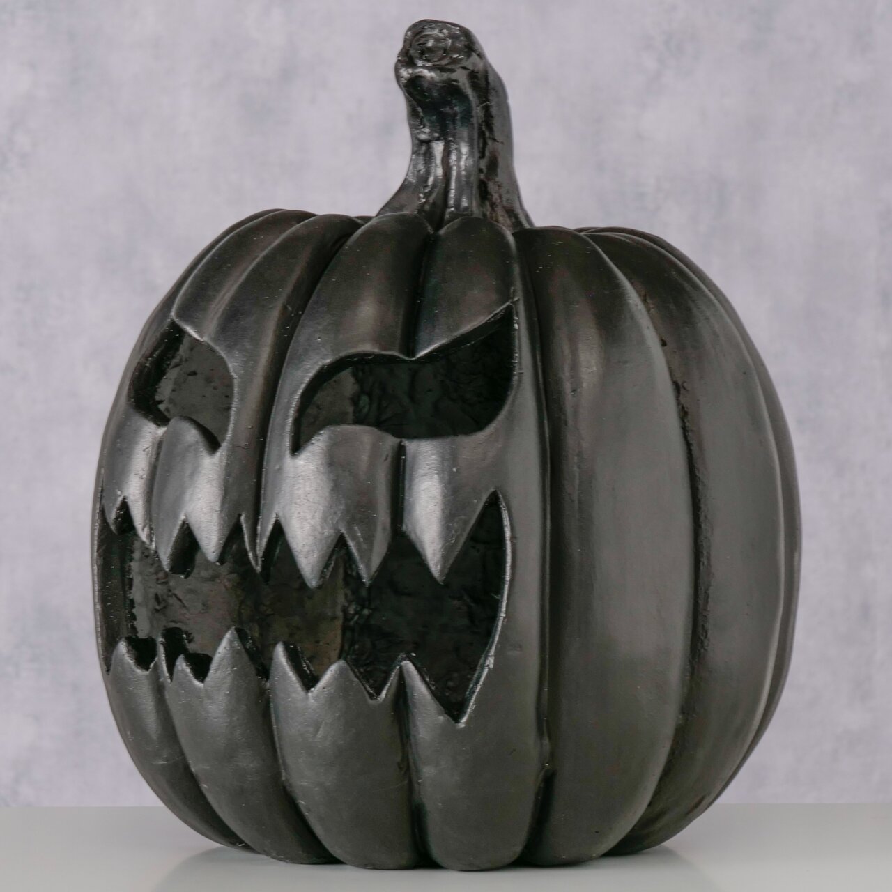 Schwarzer Halloween-Kürbis (Jack-o’-Lantern) mit grimmigem, ausgeschnittenem Gesicht und gezacktem Mund, frontal aus leicht niedriger Perspektive fotografiert vor hellem Hintergrund.