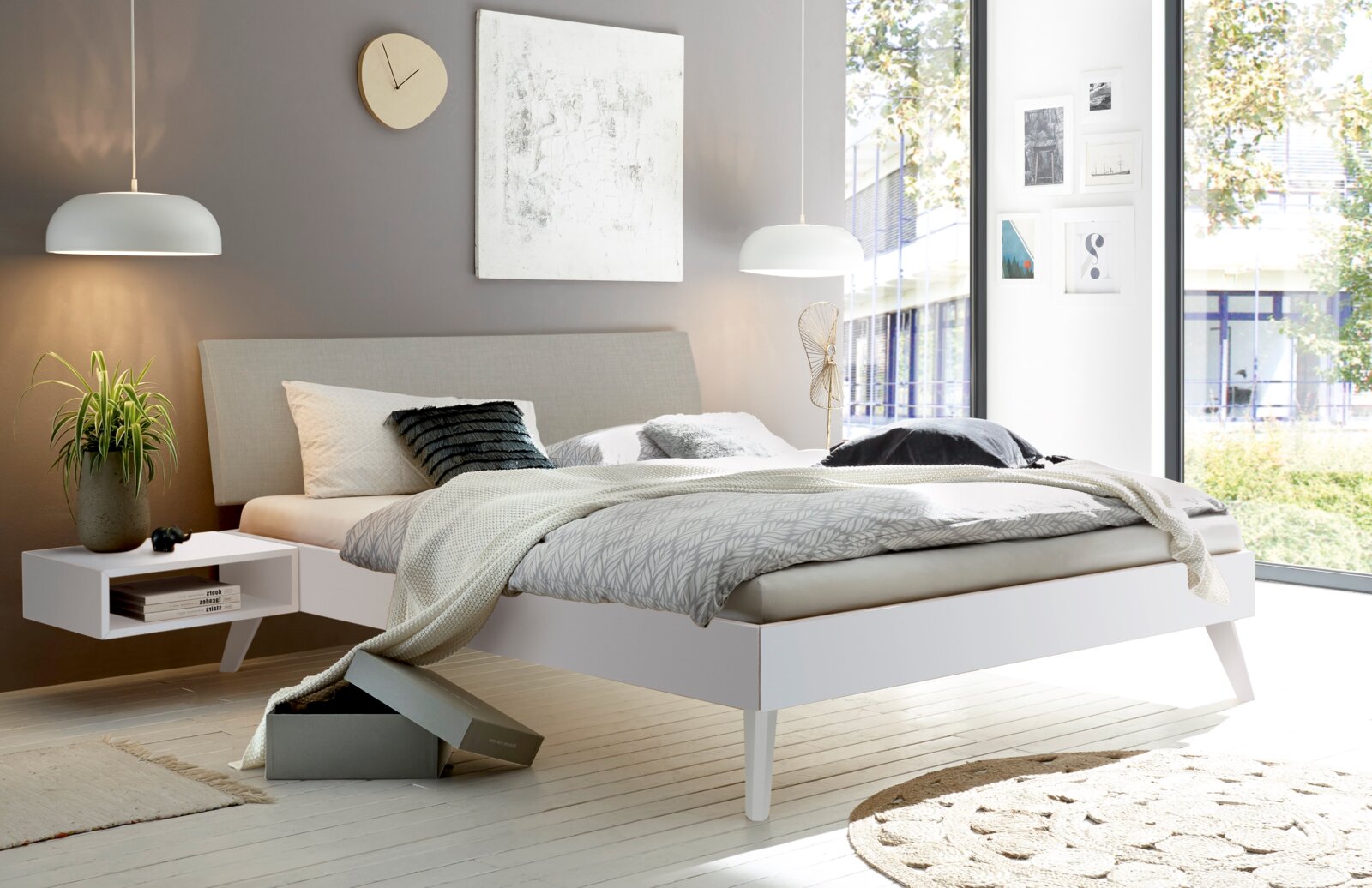 Modernes Schlafzimmer mit gepolstertem Kopfteil, seitliche Perspektive. Helles, minimalistisches Design mit hängender Lampe und Pflanzenakzent.