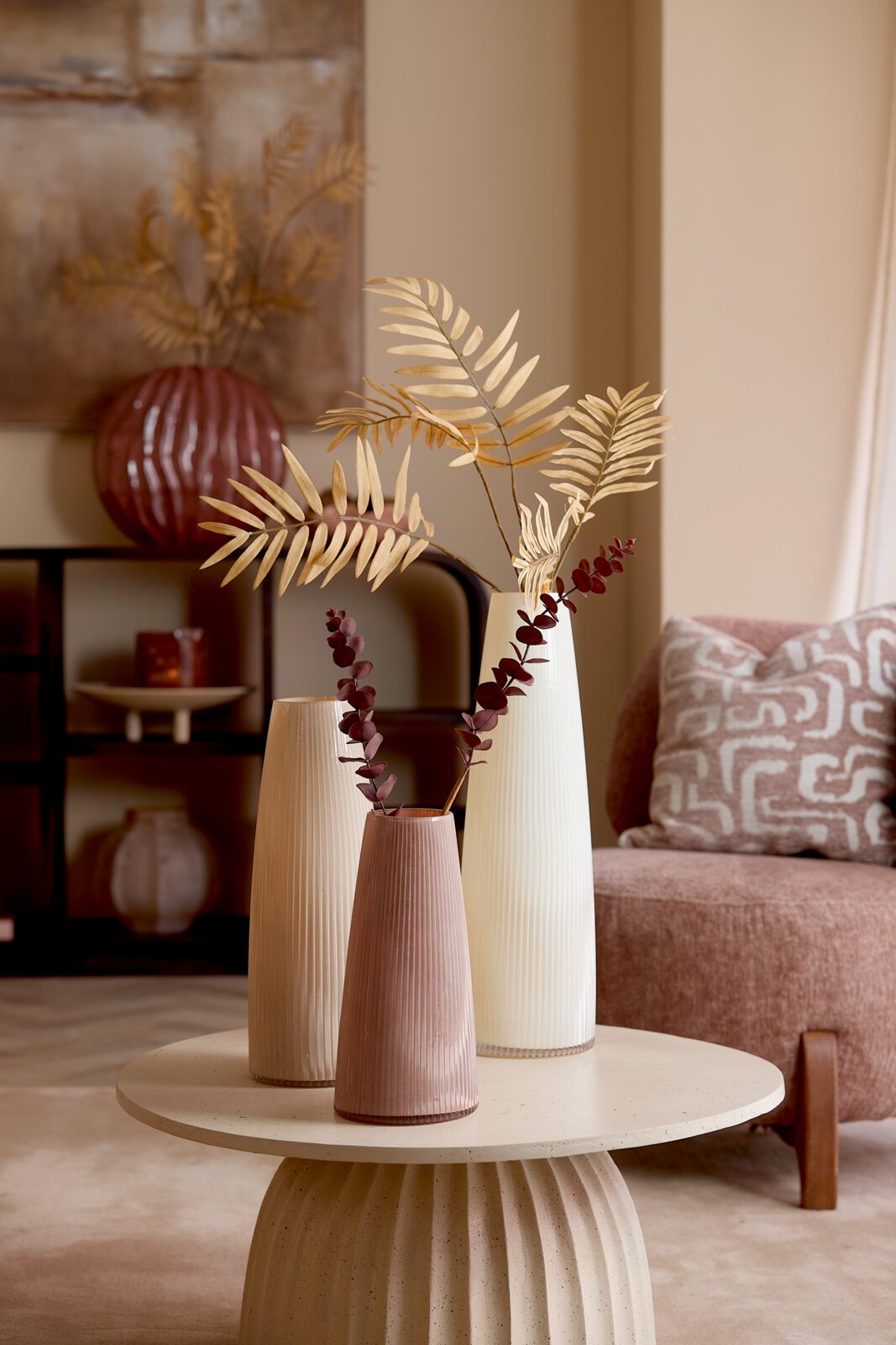 Drei geriffelte, konische Dekovasen in Creme, Beige und Altrosa mit Trockenblumen, aus leichter Frontperspektive auf einem runden Couchtisch in einem stilvoll eingerichteten Wohnzimmer.