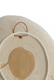 Nahaufnahme eines dekorativen Ornaments mit rissiger Textur und einem zentralen, runden Muster. Das Ornament hat eine beige und weiße Farbgebung und ist mit einer Schlaufe zum Aufhängen versehen.