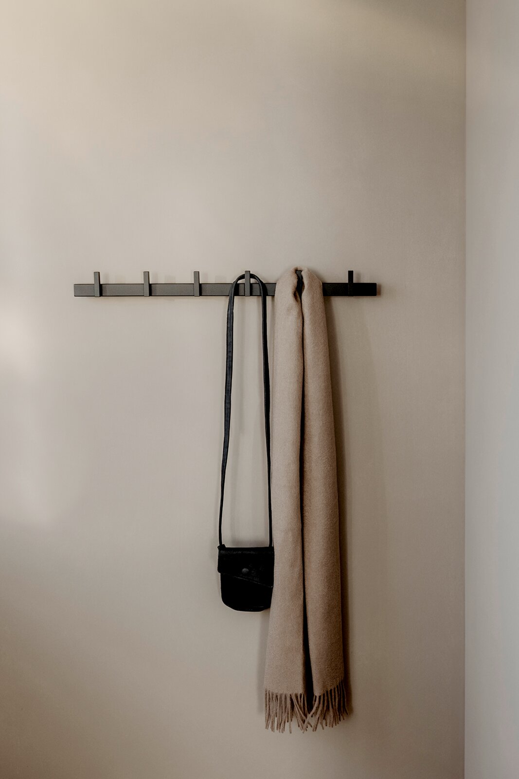 Schwarze, minimalistische Wandgarderobe mit mehreren Haken, daran hängen eine kleine schwarze Umhängetasche und ein beiger Schal; frontale Ansicht.