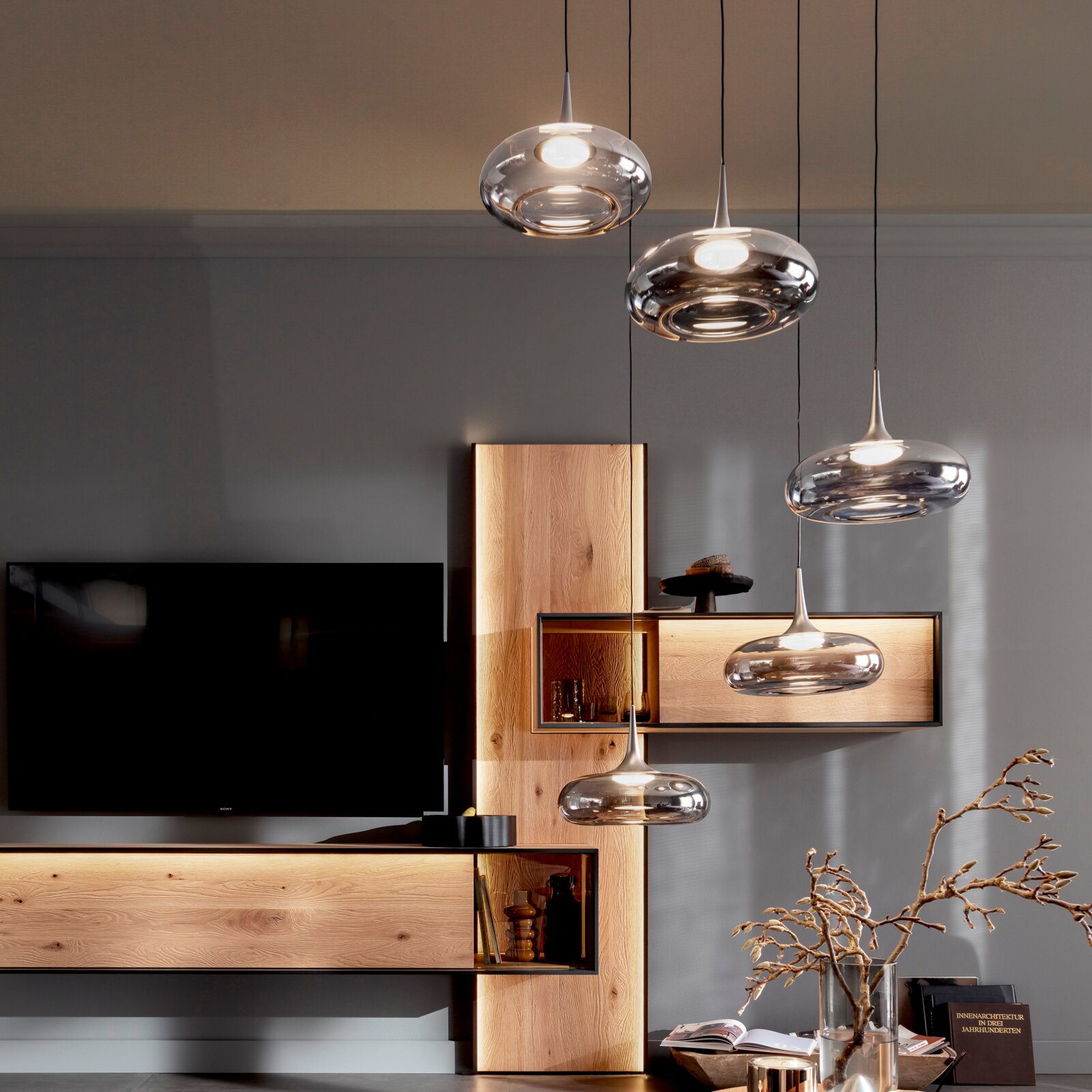 Moderne Pendelleuchten aus transparentem Glas mit warmem Licht über einem Wohnbereich mit Holzregal und Fernseher, Innenraumaufnahme in leichter Untersicht