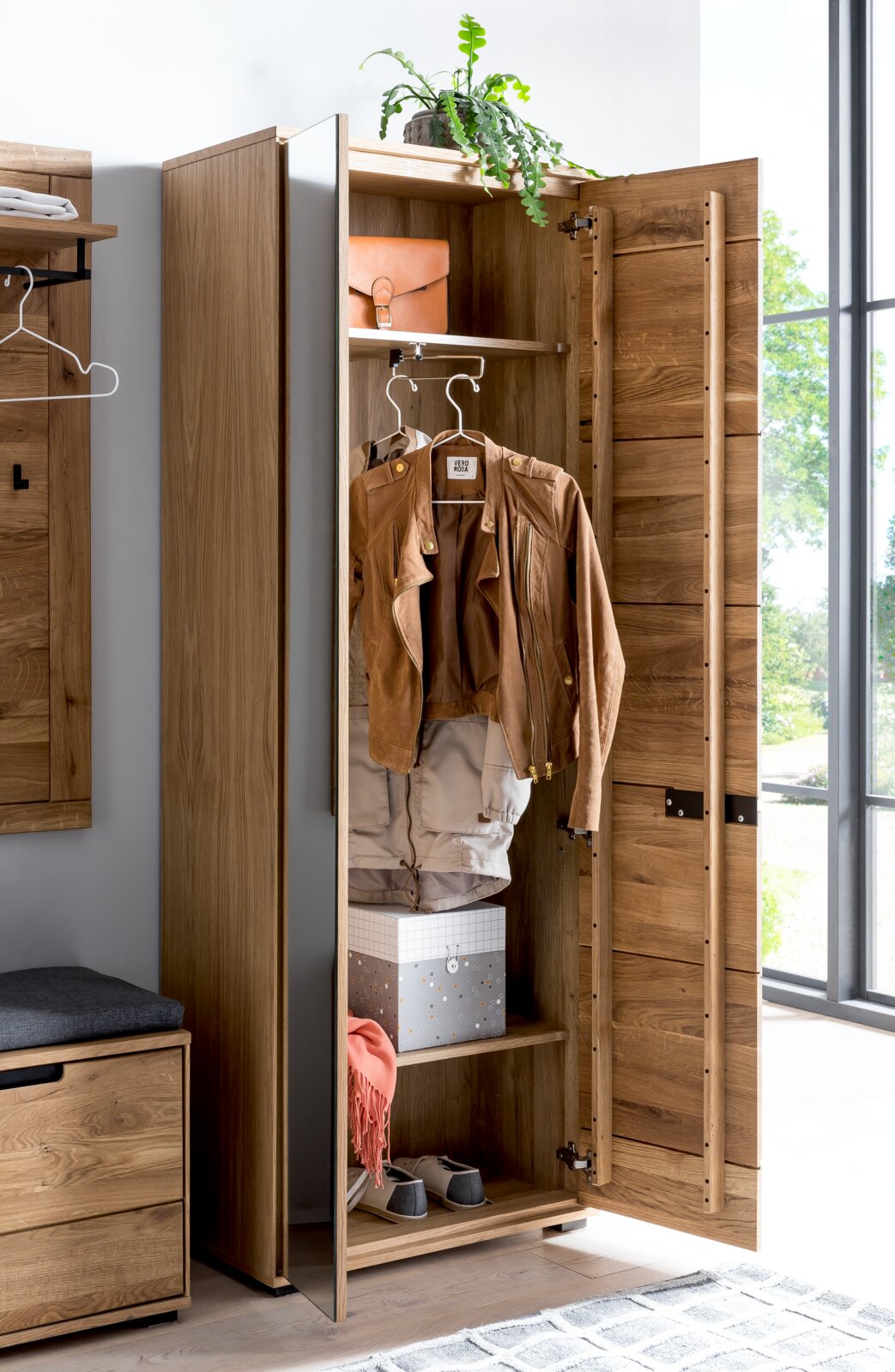 Geöffneter Garderobenschrank aus Holz mit Spiegel an der Tür, zeigt Innenraum mit Kleiderstange, Jacke, Hutablage, Schachtel und Schuhen. Perspektive von vorne.