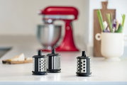 Drei verschiedene Trommelreiben in Nahaufnahme auf einer Küchenarbeitsplatte, im Hintergrund ein roter Küchenmixer.