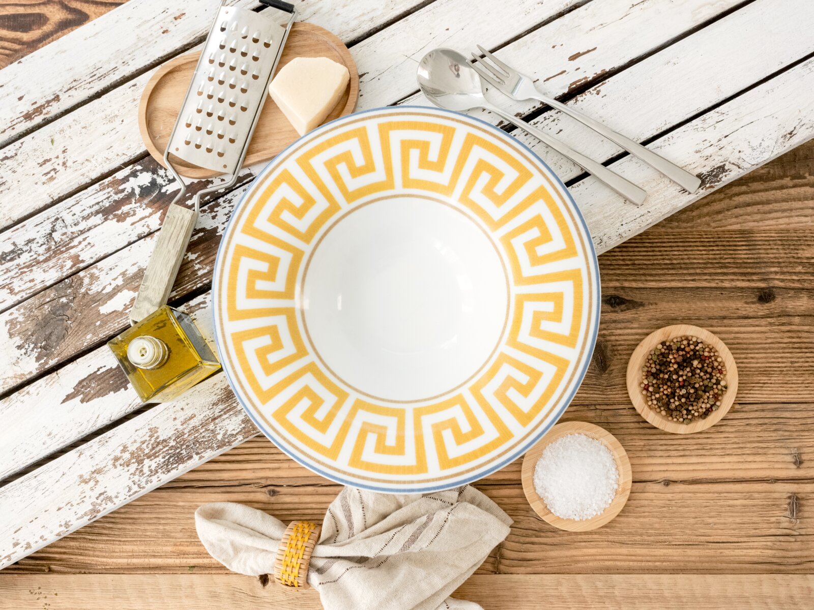 Runder Keramikteller in Weiß mit gelbem griechischem Mäander-Randmuster auf rustikalem Holztisch, umgeben von Besteck, Reibe, Käse, Öl, Salz und Pfeffer; Draufsicht von oben