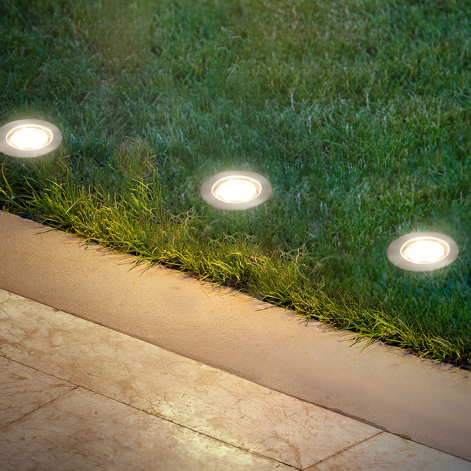 Bodenleuchten aus Edelstahl, eingebaut in eine Rasenfläche neben einem gepflasterten Weg, aus der Vogelperspektive betrachtet.