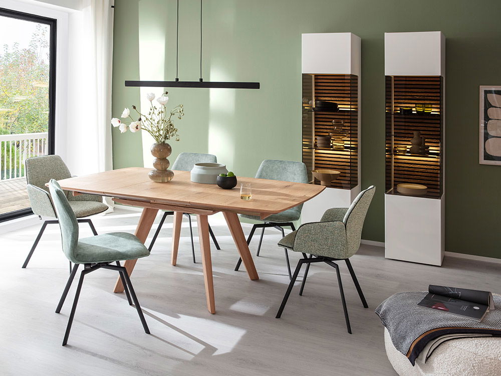 Esszimmer mit Holztisch, grünen Stühlen sowie zwei weißen Vitrinen mit Beleuchtung.