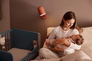 Eine Mutter hält ihr Baby in einem gemütlichen Schlafzimmer, neben einem blauen Anstellbett mit Kuscheltieren. Die Perspektive zeigt eine warme und einladende Atmosphäre.