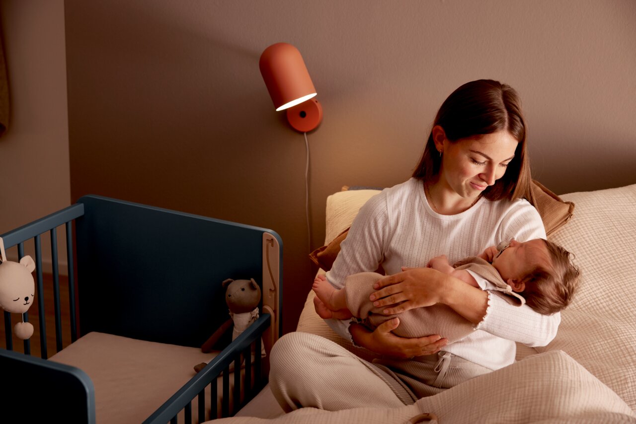 Een moeder houdt haar baby vast in een gezellige slaapkamer, naast een blauw bijzetbedje met knuffels. Het perspectief straalt een warme en uitnodigende sfeer uit.