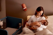 Een moeder houdt haar baby vast in een gezellige slaapkamer, naast een blauw bijzetbedje met knuffels. Het perspectief straalt een warme en uitnodigende sfeer uit.