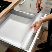 Eine Schubladenmatte wird in einer geöffneten Küchenschublade ausgerollt und mit einer Schere zugeschnitten. Perspektive von oben.