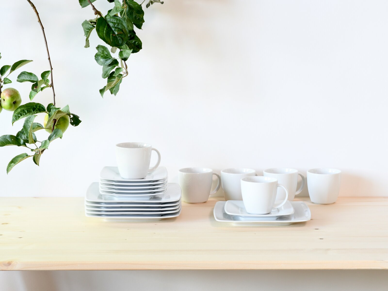 Weißes Porzellan-Kaffeeservice mit Tassen und rechteckigen Untertassen/Platten auf hellem Holztisch, links gestapelt und rechts arrangiert, mit Apfelzweig im Hintergrund; Perspektive: frontale Ansicht auf Augenhöhe.