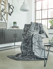 Graue Jacquard-Decke mit geometrischem Muster, drapiert über einem Sessel in einem modernen Wohnzimmer mit großen Fenstern und minimalistischer Einrichtung. Perspektive von vorne.