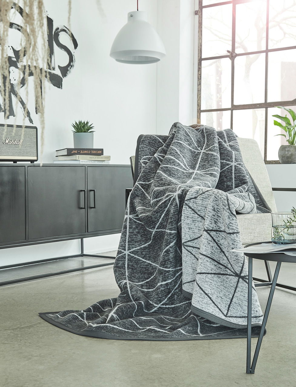 Graue Jacquard-Decke mit geometrischem Muster, drapiert über einem Sessel in einem modernen Wohnzimmer mit großen Fenstern und minimalistischer Einrichtung. Perspektive von vorne.