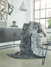 Graue Jacquard-Decke mit geometrischem Muster, drapiert über einem Sessel in einem modernen Wohnzimmer mit großen Fenstern und minimalistischer Einrichtung. Perspektive von vorne.