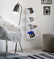 Weißes, wandmontiertes CD/DVD-Regal mit vier schrägen Ablagen, befüllt mit Medienhüllen, in modernem Wohnzimmer; Frontansicht in leichter Schrägperspektive von links.