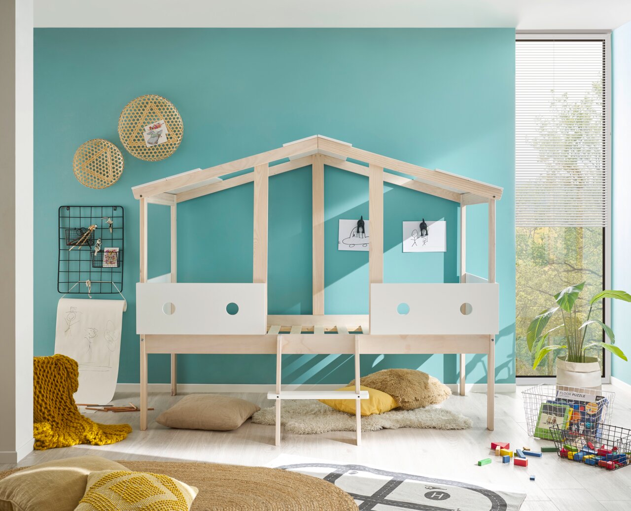 Spielbett in Hausform aus hellem Holz mit weißen Elementen, seitliche Perspektive, umgeben von bunten Kissen und Spielzeug.