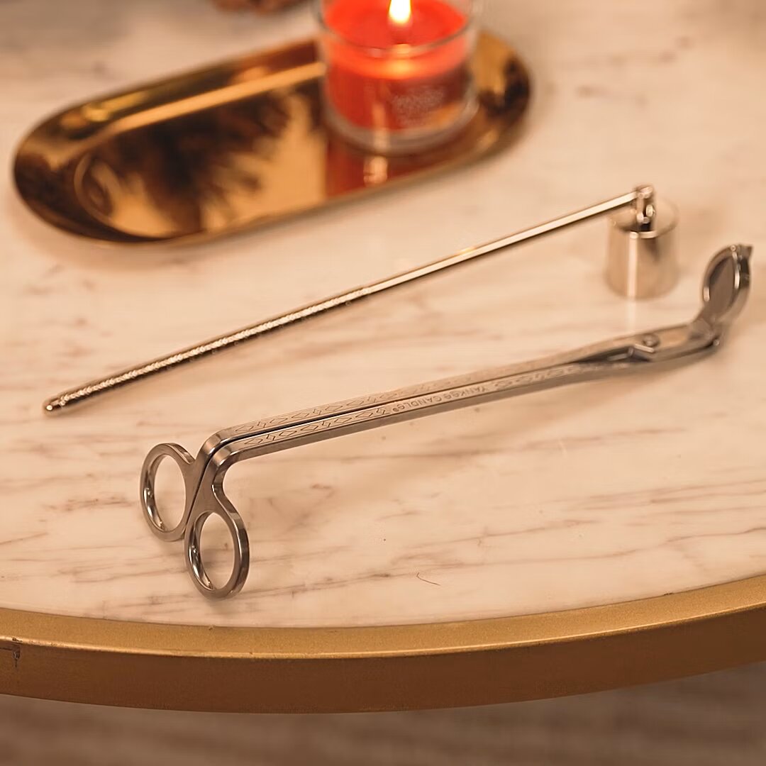 Silbernes Kerzenpflege-Set mit Dochtschere und Kerzenlöscher, aus schräger Nahperspektive auf einem eleganten Marmortisch mit brennender Duftkerze arrangiert
