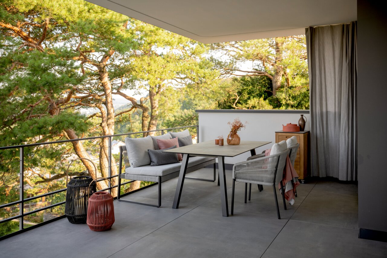 Moderner Gartentisch auf einem Balkon mit Blick auf Bäume, umgeben von stilvollen Stühlen und Dekorationen, seitliche Perspektive.
