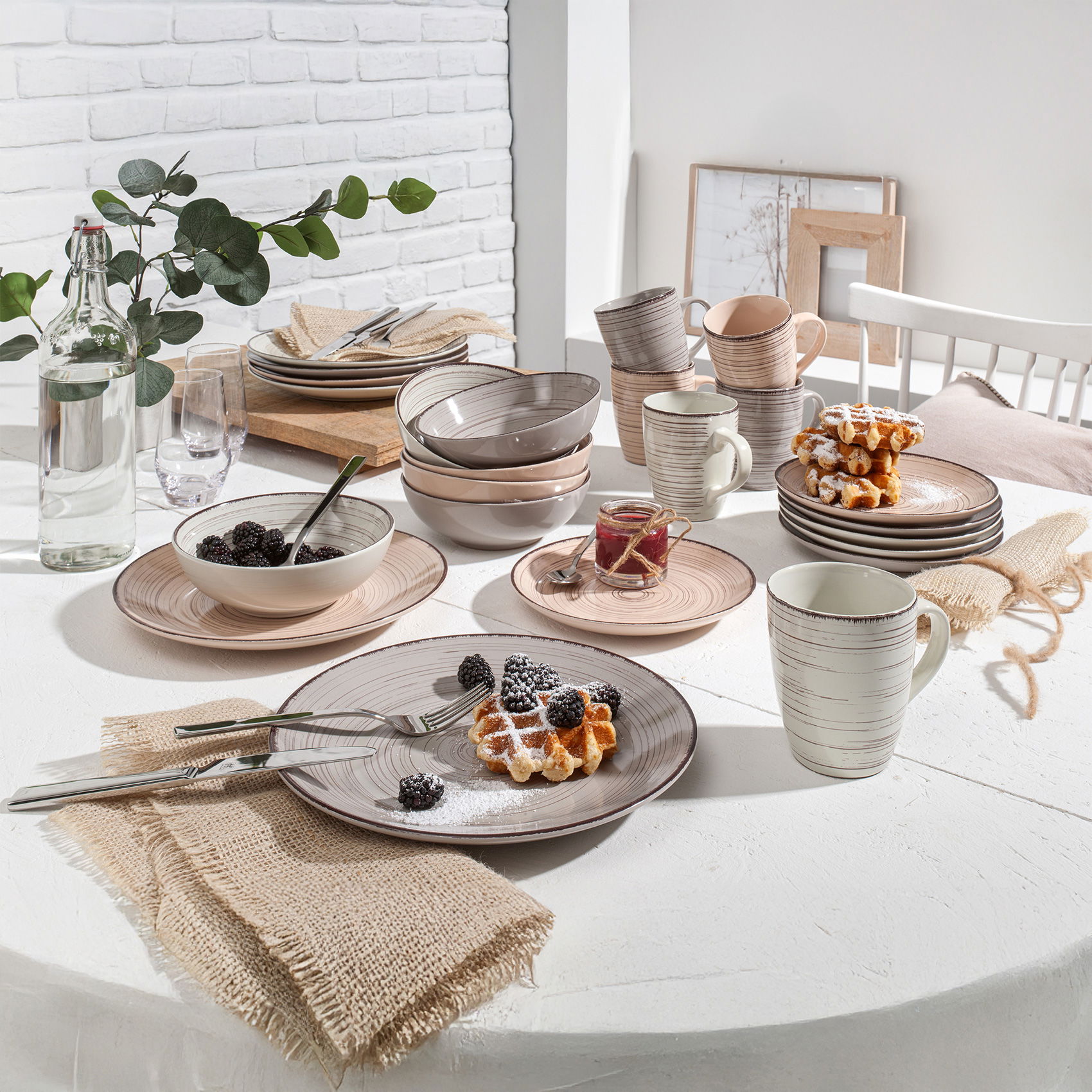 Rustikales Geschirrset mit Tellern, Schalen und Tassen, serviert mit Waffeln und Brombeeren auf hellem Tisch