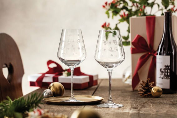 Zwei Rotweinkelche mit dekorativen Motiven auf einem festlich gedeckten Tisch, im Hintergrund Geschenke und eine Weinflasche, seitliche Perspektive.