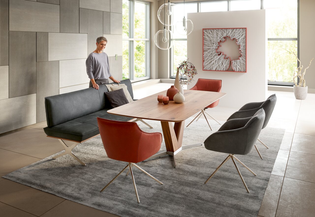 Moderner Esstisch aus Holz mit V-förmigem Gestell, umgeben von stilvollen Stühlen in einem eleganten Esszimmer mit großen Fenstern. Perspektive: seitlich.