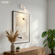 Weiße Wandspot-Leuchte mit geriffeltem Schirm und warmem Licht über einem gerahmten abstrakten Bild an heller Wand, seitliche Perspektive von links im Wohnraum mit Holz-Sideboard und Pflanzen.