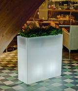 Moderner, beleuchteter Blumentopf Junco 80 in rechteckiger Form, gefüllt mit grünen Pflanzen, auf einer karierten Bodenfläche in einem Innenraum, seitliche Perspektive.